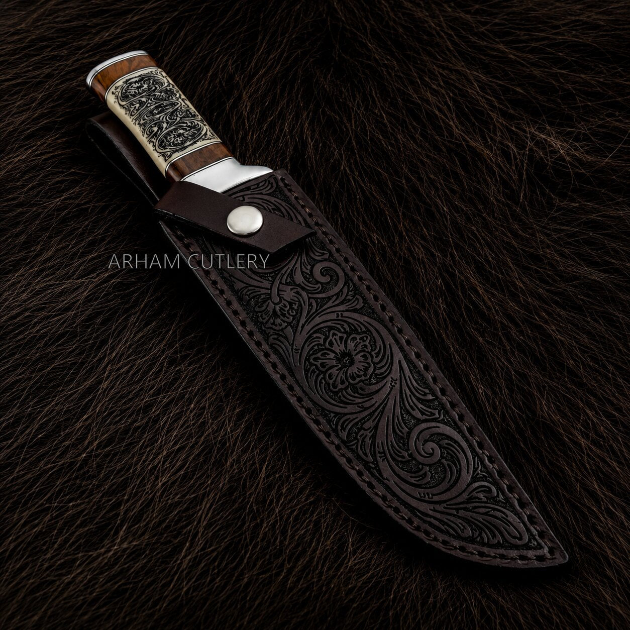An ornate fixed-blade knife with a dark brown tooled leather sheath, displayed on a dark brown fur background. The knife features a silver-toned metal bolster, a silver engraved handle scale, and a wood-toned handle.
