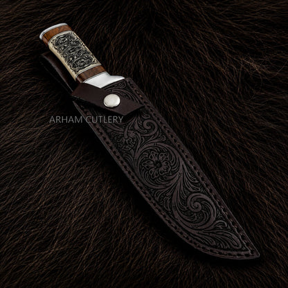 An ornate fixed-blade knife with a dark brown tooled leather sheath, displayed on a dark brown fur background. The knife features a silver-toned metal bolster, a silver engraved handle scale, and a wood-toned handle.