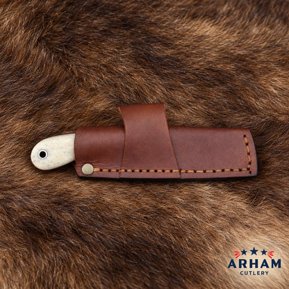 A leather knife sheath holding a knife with an off-white handle, placed on a brown fur background. The Arham Cutlery logo is visible in the bottom right corner.