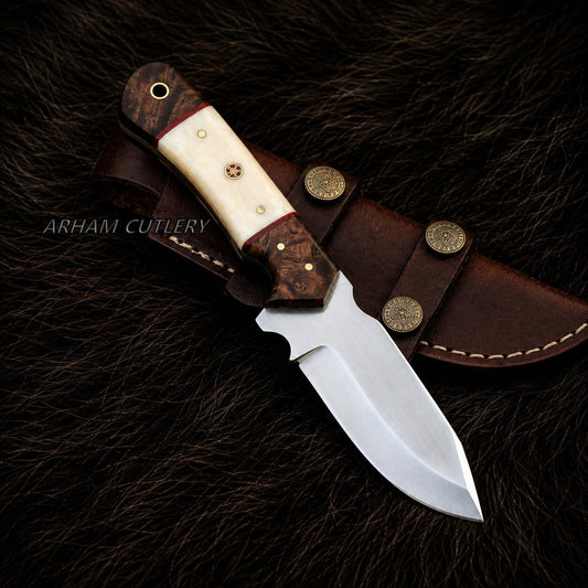 A fixed-blade knife with a polished silver blade, a cream bone handle, and a dark brown wood bolster, displayed diagonally on its brown leather sheath over a dark brown fur background. The brand name 'ARHAM CUTLERY' is watermarked on the left side.