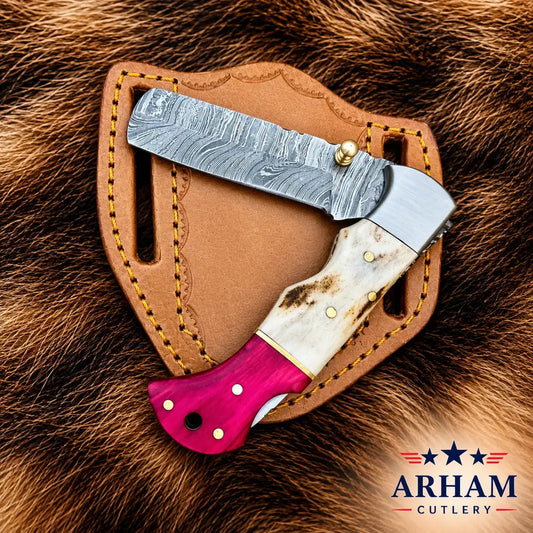 A folding knife with a Damascus steel blade, a white bone handle, and a bright pink end, displayed on a brown leather sheath over a brown fur background. The Arham Cutlery logo is visible in the bottom right corner.