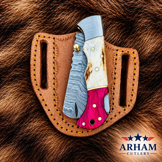 A folding pocket knife with a blue-gray Damascus steel blade, white bone handle, and bright pink inlay, displayed in a brown leather sheath on a brown fur background. The Arham Cutlery logo is visible in the bottom right corner.