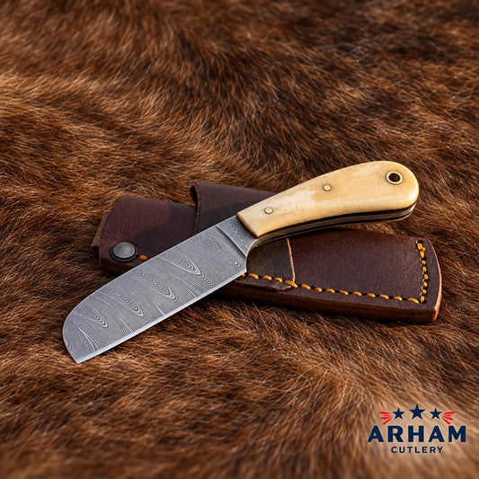 A folding knife with a Damascus steel blade and a light wood handle rests on a brown leather sheath over a brown fur background. The Arham Cutlery logo is visible in the bottom right corner.