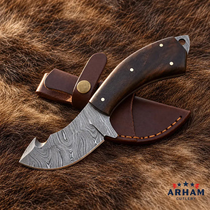 A fixed-blade knife with a patterned Damascus-style steel blade and a dark brown wooden handle, shown with a matching brown leather sheath on a brown fur background. The ARHAM CUTLERY logo appears in the bottom right corner.