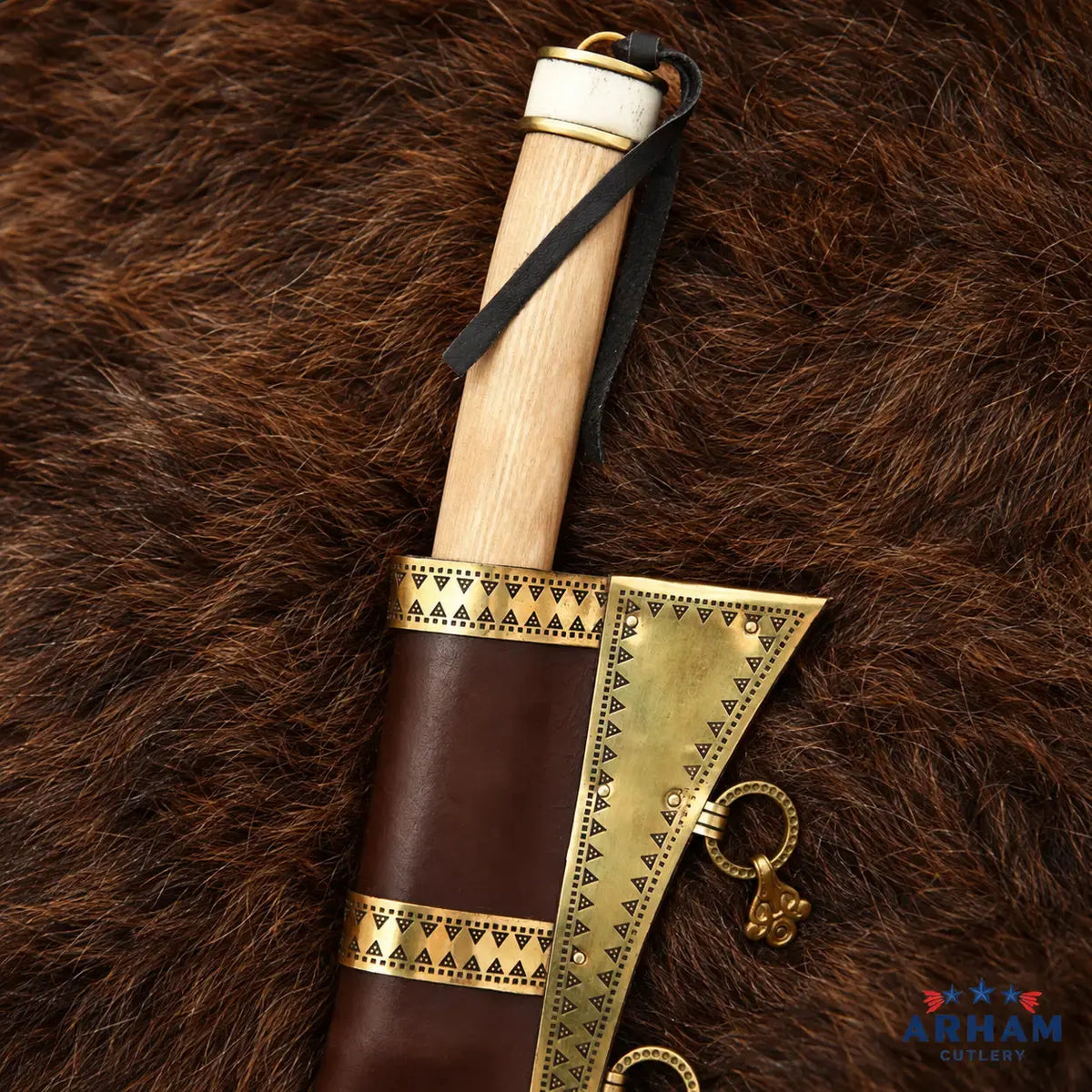 A close-up of a decorative axe or hatchet with a light wood handle, dark brown leather sheath, and ornate gold-toned metal accents, laid on a brown fur surface. The ARHAM CUTLERY logo appears in the bottom right corner.