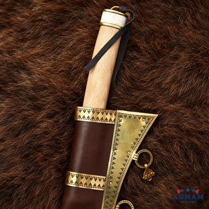A close-up of a decorative axe or hatchet with a light wood handle, dark brown leather sheath, and ornate gold-toned metal accents, laid on a brown fur surface. The ARHAM CUTLERY logo appears in the bottom right corner.