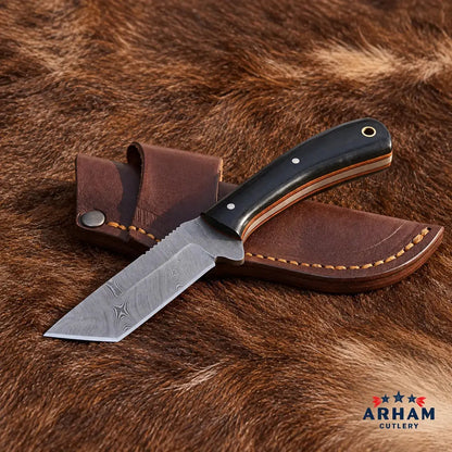 A fixed-blade knife with a dark handle and partially serrated blade, resting on a brown leather sheath over a brown fur background. The ARHAM CUTLERY logo is visible in the bottom right corner.