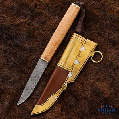 A decorative knife with a patterned steel blade, light wood handle, and brown leather sheath with ornate gold-toned metal fittings, arranged on a brown fur background. An ARHAM GILTYER logo appears in the bottom right corner.