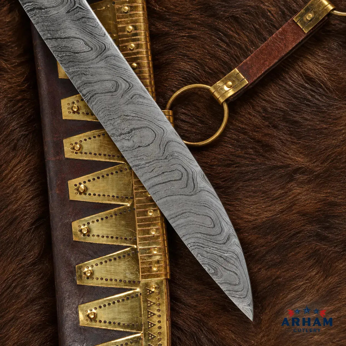 A close-up product shot of a Damascus steel knife blade laid diagonally over a dark brown leather sheath with brass-toned metal accents, placed on a brown fur surface. A small ARHAMS logo appears in the bottom right corner.