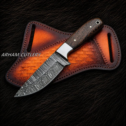 A fixed-blade knife with a patterned steel blade, a dark wood handle, and a silver bolster, laid diagonally across a brown leather sheath on a dark textured background. The text 'ARHAM CUTLERY' is visible on the left.