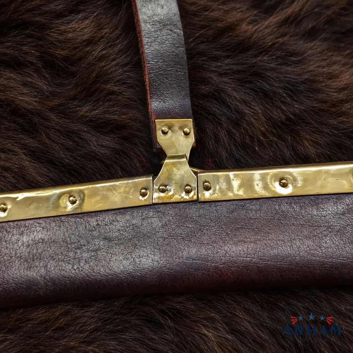 Close-up of a dark brown leather bag strap with a hammered brass clasp, resting on a dark brown fur surface. A small 'AOTAM' brand logo is visible in the bottom right corner.