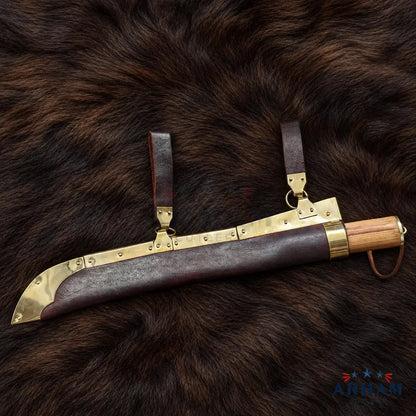 A curved machete-style knife with a dark brown leather sheath, brass metal fittings, and a light wood handle, displayed on a dark brown fur background. The ARHAM brand logo is visible in the bottom right corner.