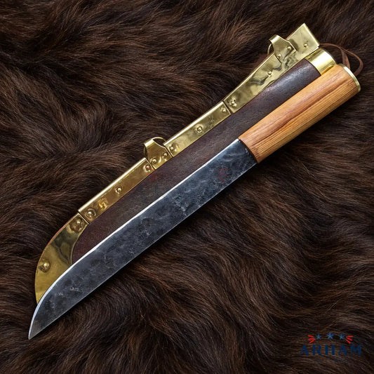 A fixed-blade knife with a dark patterned steel blade, a light wood handle, and a dark brown leather sheath with brass hardware, displayed diagonally on a dark brown fur background.