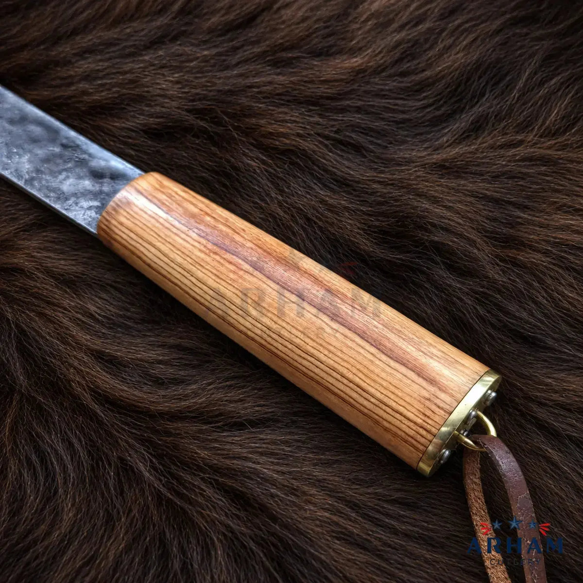 A close-up of a knife with a light brown wooden handle and a dark textured blade, resting on a dark brown fur background. A small leather loop with a brass ring is attached to the handle.