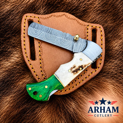 A folding pocket knife with a Damascus steel blade, white bone handle, and green contoured handle, resting on its brown leather sheath over a brown fur background. The Arham Cutlery logo is visible in the bottom right corner.