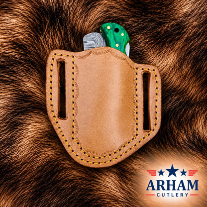 A tan leather knife sheath with yellow stitching, holding a folding knife with a green handle, set against a brown fur background. The Arham Cutlery logo is visible in the bottom right corner.