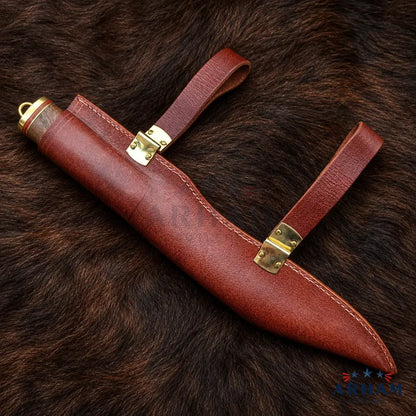 A brown leather sheath with brass hardware and two straps, laid diagonally on a dark brown fur background. A faint ARTI watermark is visible across the center, and an ARTI logo appears in the bottom right corner.