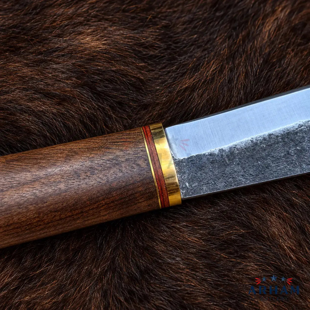 A close-up of a knife with a dark brown wooden handle and a patterned steel blade, resting on a brown fur background. The handle is accented with gold-colored metal bands.