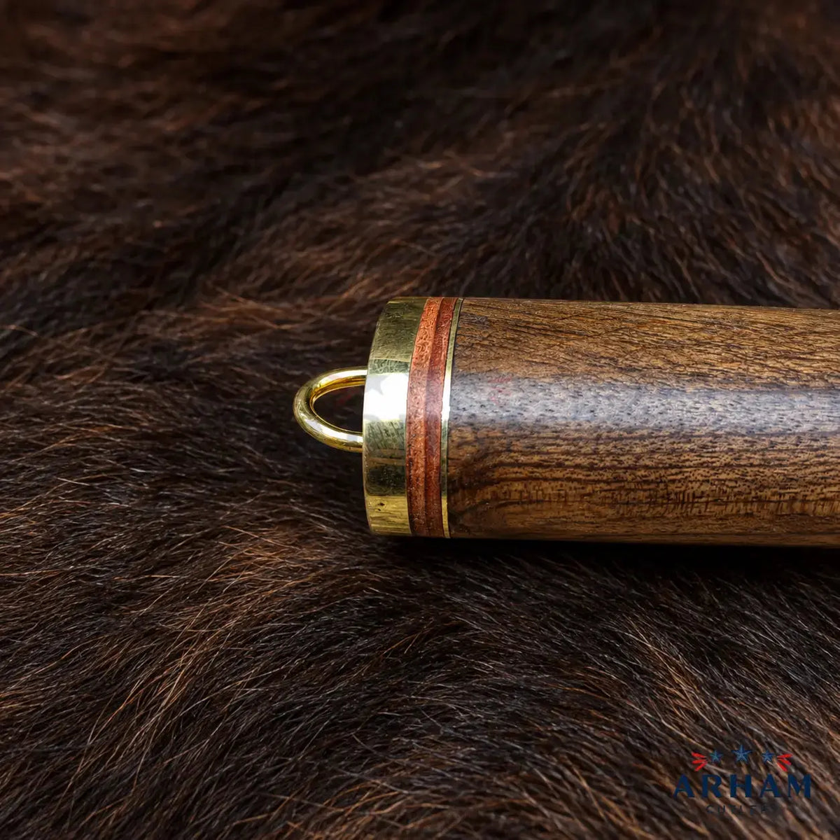 Close-up of a wooden cylindrical product with a brass ring and loop, resting on dark brown animal fur. The wood has a prominent grain pattern with a reddish-brown inlay stripe.