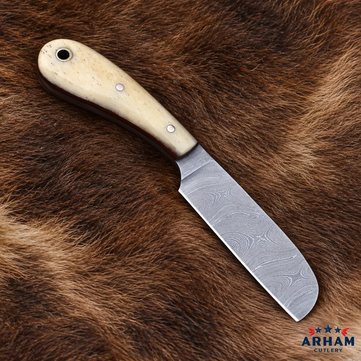 A fixed-blade knife with a patterned Damascus steel blade and a cream-colored bone handle, displayed diagonally on a brown animal fur background. The Arham Cutlery logo is visible in the bottom right corner.