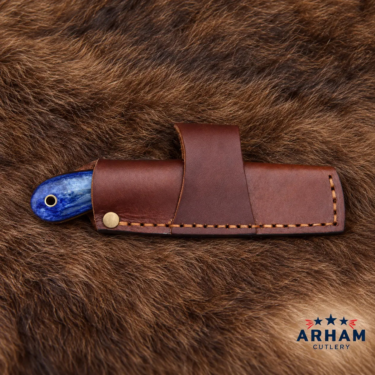 A brown leather knife sheath with a brass snap and contrast stitching, holding a knife with a blue marbled handle, laid on a brown fur background. The ARHAM CUTLERY logo is visible in the bottom right corner.
