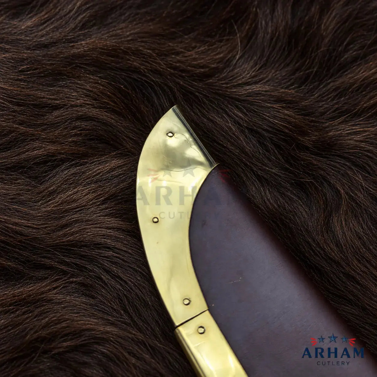 Close-up of a curved brass knife blade with rivets, resting on dark brown fur. A dark brown leather or wood handle is partially visible, with ARHAM CUTLERY branding shown on the blade and in the bottom right corner.