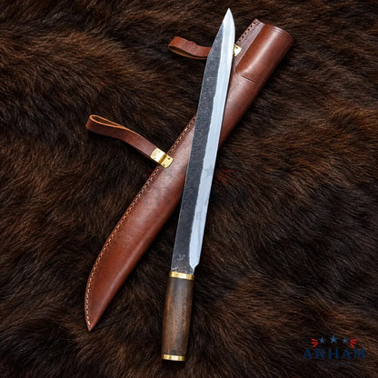 A fixed-blade knife with a textured steel blade, dark wood handle, and brass accents, displayed diagonally next to its brown leather sheath on a dark brown fur background. The ARHAM brand logo is visible in the bottom right corner.