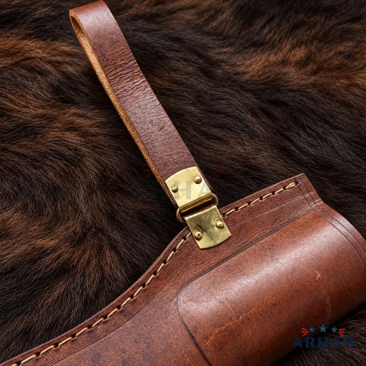 A close-up of a brown leather holster with a matching leather strap and brass buckle, laid on dark brown fur. A small AR-15 logo watermark is visible in the bottom right corner.