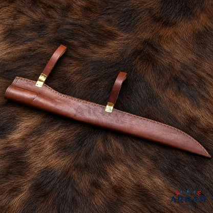 A brown leather knife sheath with brass-colored metal accents, laid diagonally on a dark brown animal fur background.