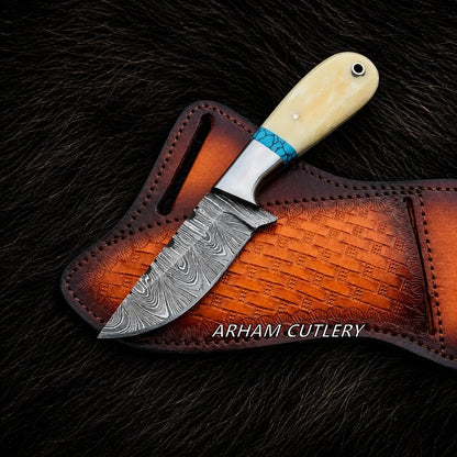 A fixed-blade knife with a patterned Damascus steel blade, a turquoise and silver wrapped bolster, and a cream-colored bone handle, resting on a brown embossed leather sheath over a dark fur background. The brand name 'ARHAM CUTLERY' is printed on the sheath.