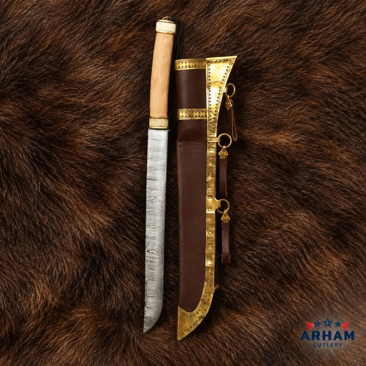 A custom hand-forged knife with a textured steel blade, a light wood handle, and brass fittings, displayed next to its brown leather sheath with brass trim, laid flat on a brown fur background. The Arham Cutlery logo is visible in the bottom right corner.