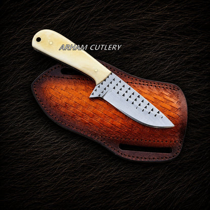A fixed-blade knife with a cream-colored bone handle and a patterned silver blade, resting on a brown leather sheath over a dark textured fur background. The text 'ARHAM CUTLERY' is printed on the blade.