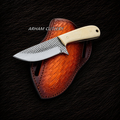 A fixed-blade knife with a patterned steel blade and cream-colored handle, resting on a brown leather sheath over a dark textured background. The text 'ARHAM CUTLERY' is overlaid on the image.