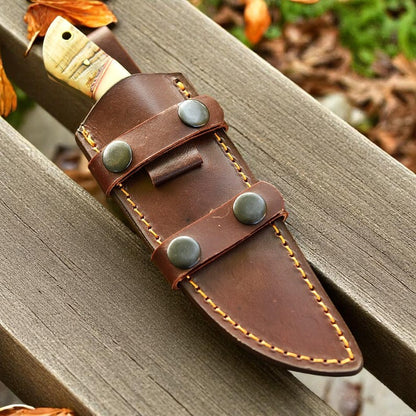 D2 steel knife with ram horn and camel bone handle in leather sheath on wooden bench.

