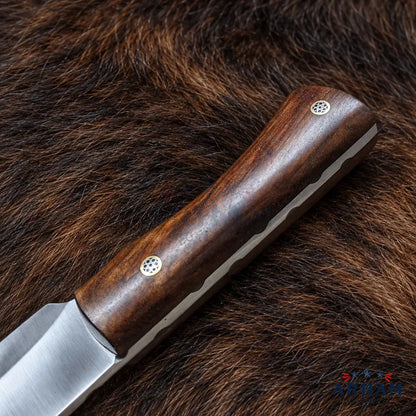 A close-up of a knife with a dark brown wooden handle and a satin-finished steel blade, placed diagonally on a brown fur surface. The handle has two small circular metal rivets.
