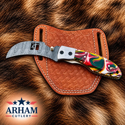 A folding pocket knife with a patterned steel blade and a colorful marbled handle, resting on a brown leather sheath over a brown fur background. The ARHAM CUTLERY logo is visible in the bottom-left corner.