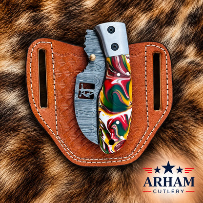 A folding pocket knife with a marbled multicolor handle and a textured gray metal blade, displayed in a brown leather sheath on a brown fur background. The Arham Cutlery logo is visible in the bottom right corner.