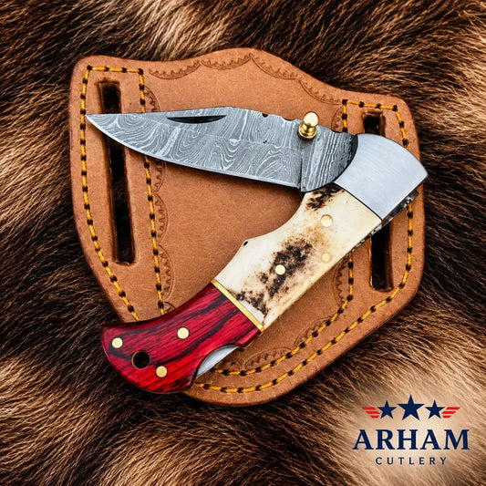 A folding pocket knife with a patterned steel blade, a cream-colored bone handle, and a red marbled wood bolster, displayed on a brown leather sheath over a fur background. The ARHAM CUTLERY logo is visible in the bottom right corner.