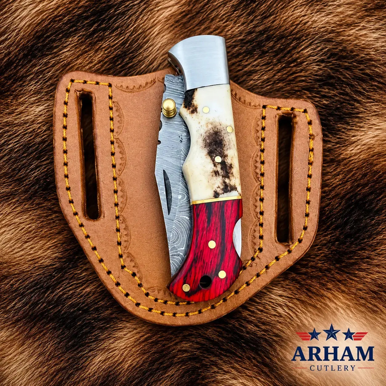A folding pocket knife with a red and white wood handle and Damascus steel blade, tucked into a brown leather sheath with gold stitching, placed on a brown fur background. The ARHAM CUTLERY logo is visible in the bottom right corner.