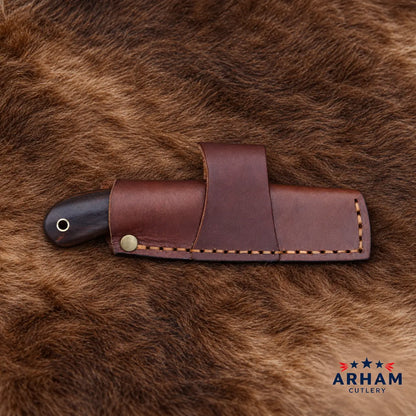 A brown leather knife sheath holding a knife with a dark handle, laid on a brown fur surface. The ARHAM CUTLERY logo appears in the bottom right corner.