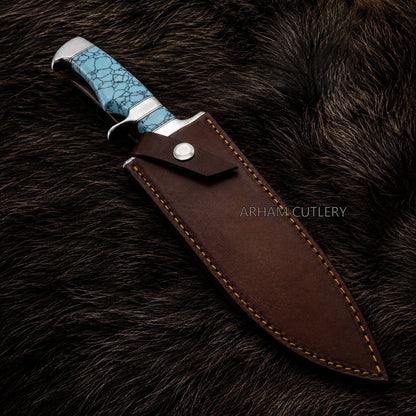 A fixed-blade knife with a textured turquoise blue handle and silver bolster, shown partially sheathed in a dark brown leather sheath with tan stitching, placed on a dark brown fur background. The brand name 'ARHAM CUTLERY' is watermarked on the sheath.