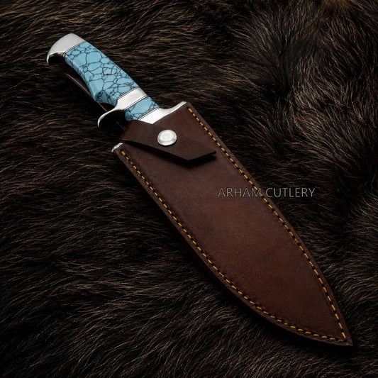A fixed-blade knife with a textured turquoise blue handle and silver bolster, shown partially sheathed in a dark brown leather sheath with tan stitching, placed on a dark brown fur background. The brand name 'ARHAM CUTLERY' is watermarked on the sheath.