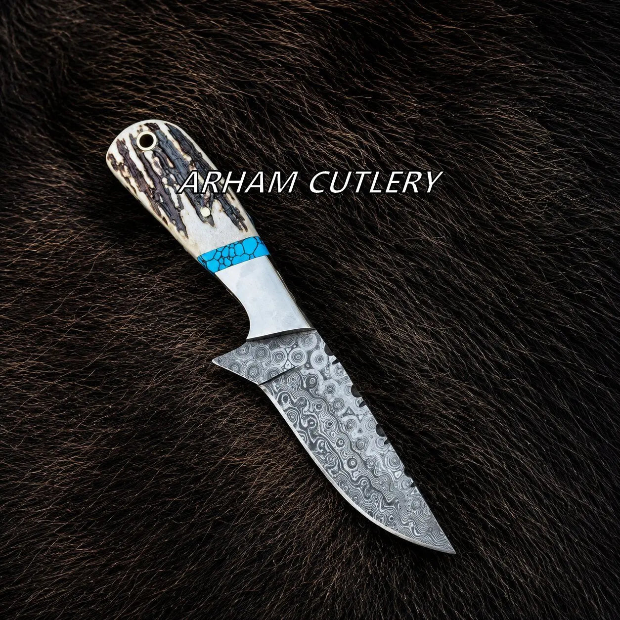 A fixed-blade knife with a patterned steel blade and a textured handle featuring a bright blue accent band, displayed diagonally on a dark brown fur background. The text 'ARHAM CUTLERY' is overlaid across the handle.