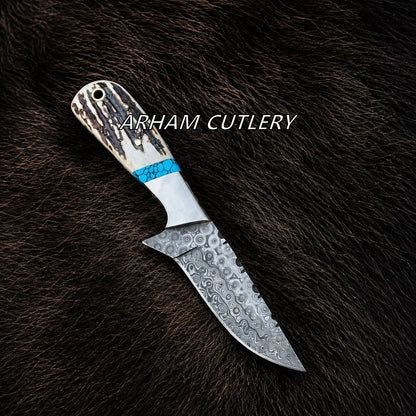 A fixed-blade knife with a patterned steel blade and a textured handle featuring a bright blue accent band, displayed diagonally on a dark brown fur background. The text 'ARHAM CUTLERY' is overlaid across the handle.