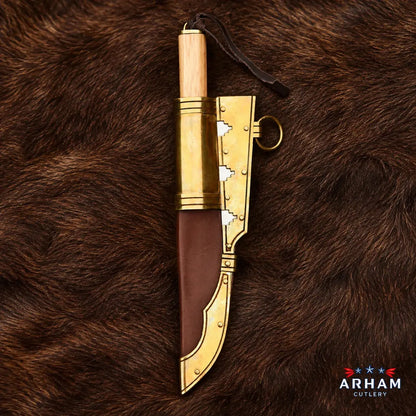 A decorative cutlass-style knife with a light wood handle, gold-toned metal guard and blade accents, and a brown leather sheath, displayed on a brown fur background. The ARHAM CUTLERY logo is visible in the bottom right corner.