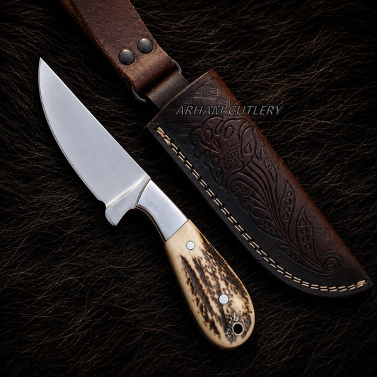 A fixed-blade knife with a polished silver blade, a silver bolster, and a carved bone handle with dark brown veining, displayed next to its dark brown embossed leather sheath on a dark textured background. A watermark for 'ARHANT CUTLERY' is visible.