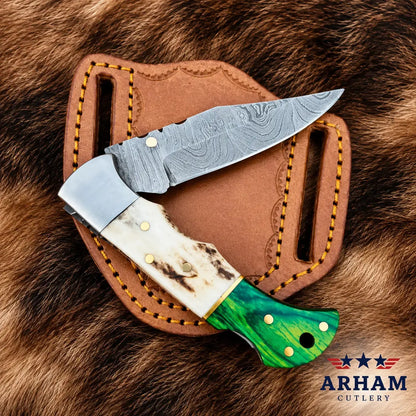 A folding pocket knife with a Damascus steel blade, bone handle, and green marbled handle scale, displayed on top of a brown leather sheath over a brown fur background. The ARHAM CUTLERY logo is visible in the bottom right corner.