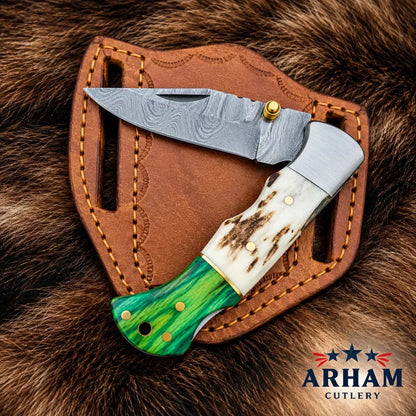 A folding pocket knife with a patterned steel blade, a white bone handle, and a green inlay handle, displayed on a brown leather sheath over a brown fur background. The ARHAM CUTLERY logo is visible in the bottom right corner.