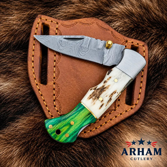 A folding pocket knife with a patterned steel blade, a white bone handle, and a green inlay handle, displayed on a brown leather sheath over a brown fur background. The ARHAM CUTLERY logo is visible in the bottom right corner.