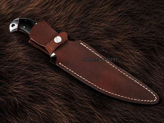 A fixed-blade knife with a dark handle, shown partially sheathed in a brown leather sheath with white stitching, placed on a dark brown fur background. The brand name 'ARSHAM CUTLERY' is printed on the leather sheath.